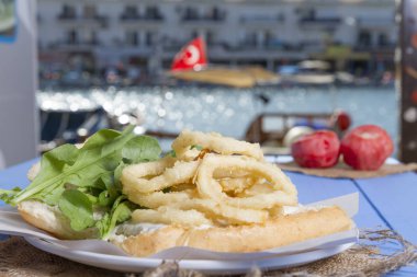 Lezzetli derin kızarmış halkalar sandviç hizmet beyaz daire plaka, yarım ekmek, soğan ve yeşil roka salatası rustik mavi ahşap arka plan üzerine dilimlenmiş. Kalamar çok mutfaklarının yenir. Üstten görünüm.