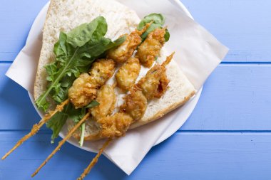 Lezzetli altın derin kızarmış pirzola midye şişleri yoğurt sos tartar ile hizmet veren sandviç, domates, dilimlenmiş beyaz soğan, maydanoz ve yeşil roka salatası mavi rustik masa arka plan üzerinde. Midye tava ekmek.