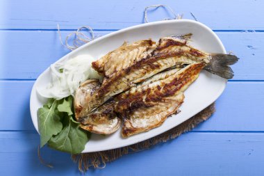 Lezzetli ızgara fileto levrek porsiyon dilimlenmiş soğan, domates, yeşil roka salatası ve sulu limon rustik mavi ahşap masa arka plan üzerinde oval beyaz plakalı füme. Alan, kopyalama kimse.