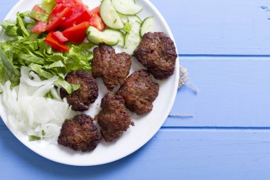 Izgara sığır eti köfte porsiyon dilimlenmiş salatalık, beyaz soğan, küp daire plakalı domates ve marul salatası mavi rustik ahşap masanın üstüne kesmek. Ekmek kırıntıları, tereyağı ve baharat gibi maddeler.