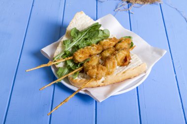 Lezzetli altın derin kızarmış pirzola midye şişleri yoğurt sos tartar ile hizmet veren sandviç, domates, dilimlenmiş beyaz soğan, maydanoz ve yeşil roka salatası mavi rustik masa arka plan üzerinde. Midye tava ekmek.