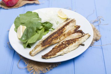 Lezzetli ızgara fileto levrek porsiyon dilimlenmiş soğan, domates, yeşil roka salatası ve sulu limon rustik mavi ahşap masa arka plan üzerinde oval beyaz plakalı füme. Alan, kopyalama kimse.