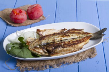 Lezzetli ızgara fileto levrek porsiyon dilimlenmiş soğan, domates, yeşil roka salatası ve sulu limon rustik mavi ahşap masa arka plan üzerinde oval beyaz plakalı füme. Alan, kopyalama kimse.