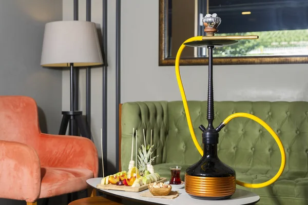 Traditional Arabic and Turkish shisha, hookah or Water pipe serving with fresh fruits, hot black tea and nuts on table, modern cafe lounge design. Led light in water.
