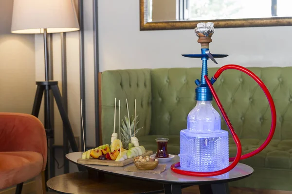 Traditional Arabic and Turkish shisha, hookah or Water pipe serving with fresh fruits, hot black tea and nuts on table, modern cafe lounge design. Led light in water.