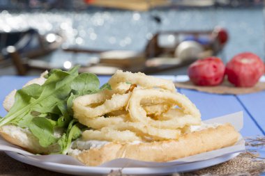 Lezzetli derin kızarmış halkalar sandviç hizmet beyaz daire plaka, yarım ekmek, soğan ve yeşil roka salatası rustik mavi ahşap arka plan üzerine dilimlenmiş. Kalamar çok mutfaklarının yenir. Üstten görünüm.