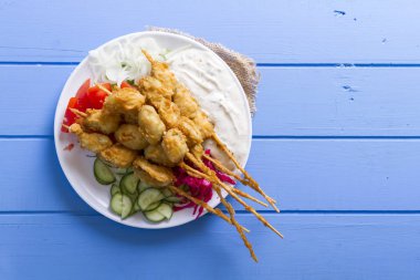 Lezzetli altın derin kızarmış pirzola midye yoğurt sos tartar ile hizmet veren şişleri, domates, dilimlenmiş beyaz soğan, maydanoz ve yeşil roka salatası mavi rustik masa arka plan üzerinde. Midye tava.