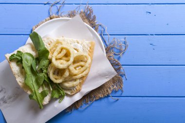 Lezzetli derin kızarmış halkalar sandviç hizmet beyaz daire plaka, yarım ekmek, soğan ve yeşil roka salatası rustik mavi ahşap arka plan üzerine dilimlenmiş. Kalamar çok mutfaklarının yenir. Üstten görünüm.