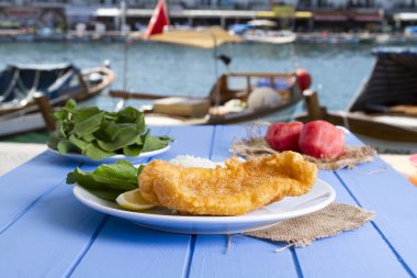 Lezzetli altın derin kızarmış mezgit balığı yeşil roka salatası ile hizmet veren fileto dilimlenmiş beyaz soğan, domates ve sulu limon yuvarlak tabakta, rustik mavi tablo arka planı. Kopya alanı için metin alanı.