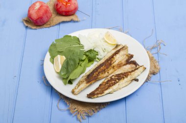 Lezzetli ızgara fileto levrek porsiyon dilimlenmiş soğan, domates, yeşil roka salatası ve sulu limon rustik mavi ahşap masa arka plan üzerinde oval beyaz plakalı füme. Alan, kopyalama kimse.