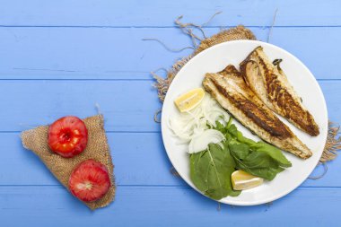 Lezzetli ızgara fileto levrek porsiyon dilimlenmiş soğan, domates, yeşil roka salatası ve sulu limon rustik mavi ahşap masa arka plan üzerinde oval beyaz plakalı füme. Alan, kopyalama kimse.