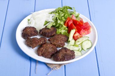 Izgara sığır eti köfte porsiyon dilimlenmiş salatalık, beyaz soğan, küp daire plakalı domates ve marul salatası mavi rustik ahşap masanın üstüne kesmek. Ekmek kırıntıları, tereyağı ve baharat gibi maddeler.