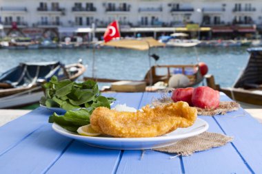 Lezzetli altın derin kızarmış mezgit balığı yeşil roka salatası ile hizmet veren fileto dilimlenmiş beyaz soğan, domates ve sulu limon yuvarlak tabakta, rustik mavi tablo arka planı. Kopya alanı için metin alanı.