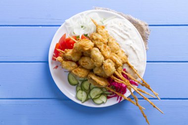 Lezzetli altın derin kızarmış pirzola midye yoğurt sos tartar ile hizmet veren şişleri, domates, dilimlenmiş beyaz soğan, maydanoz ve yeşil roka salatası mavi rustik masa arka plan üzerinde. Midye tava.