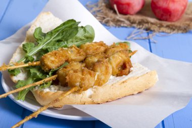 Lezzetli altın derin kızarmış pirzola midye şişleri yoğurt sos tartar ile hizmet veren sandviç, domates, dilimlenmiş beyaz soğan, maydanoz ve yeşil roka salatası mavi rustik masa arka plan üzerinde. Midye tava ekmek.