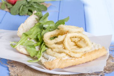 Lezzetli derin kızarmış halkalar sandviç hizmet beyaz daire plaka, yarım ekmek, soğan ve yeşil roka salatası rustik mavi ahşap arka plan üzerine dilimlenmiş. Kalamar çok mutfaklarının yenir. Üstten görünüm.