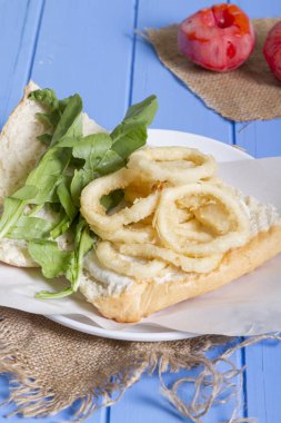 Lezzetli derin kızarmış halkalar sandviç hizmet beyaz daire plaka, yarım ekmek, soğan ve yeşil roka salatası rustik mavi ahşap arka plan üzerine dilimlenmiş. Kalamar çok mutfaklarının yenir. Üstten görünüm.