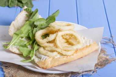 Lezzetli derin kızarmış halkalar sandviç hizmet beyaz daire plaka, yarım ekmek, soğan ve yeşil roka salatası rustik mavi ahşap arka plan üzerine dilimlenmiş. Kalamar çok mutfaklarının yenir. Üstten görünüm.