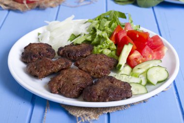 Izgara sığır eti köfte porsiyon dilimlenmiş salatalık, beyaz soğan, küp daire plakalı domates ve marul salatası mavi rustik ahşap masanın üstüne kesmek. Ekmek kırıntıları, tereyağı ve baharat gibi maddeler.