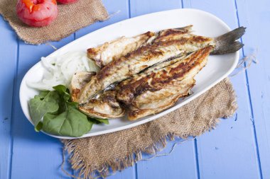 Lezzetli ızgara fileto levrek porsiyon dilimlenmiş soğan, domates, yeşil roka salatası ve sulu limon rustik mavi ahşap masa arka plan üzerinde oval beyaz plakalı füme. Alan, kopyalama kimse.