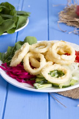 Lezzetli derin kızarmış kalamar garson beyaz daire plaka, kırmızı lahana, tartar, halka dilimlenmiş soğan ve yeşil roka salatası rustik mavi ahşap arka plan üzerinde. Kalamar çok mutfaklarının yemek. Üstten görünüm.