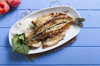 Lezzetli ızgara fileto levrek porsiyon dilimlenmiş soğan, domates, yeşil roka salatası ve sulu limon rustik mavi ahşap masa arka plan üzerinde oval beyaz plakalı füme. Alan, kopyalama kimse.