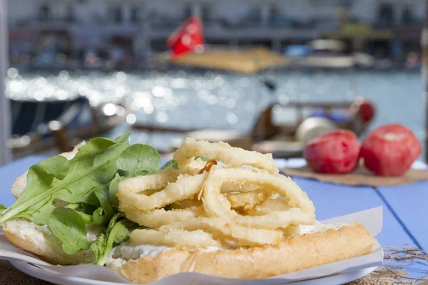 Lezzetli derin kızarmış halkalar sandviç hizmet beyaz daire plaka, yarım ekmek, soğan ve yeşil roka salatası rustik mavi ahşap arka plan üzerine dilimlenmiş. Kalamar çok mutfaklarının yenir. Üstten görünüm.