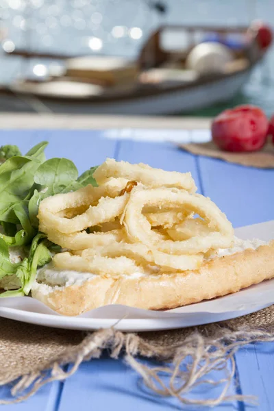 Lezzetli derin kızarmış halkalar sandviç hizmet beyaz daire plaka, yarım ekmek, soğan ve yeşil roka salatası rustik mavi ahşap arka plan üzerine dilimlenmiş. Kalamar çok mutfaklarının yenir. Üstten görünüm.