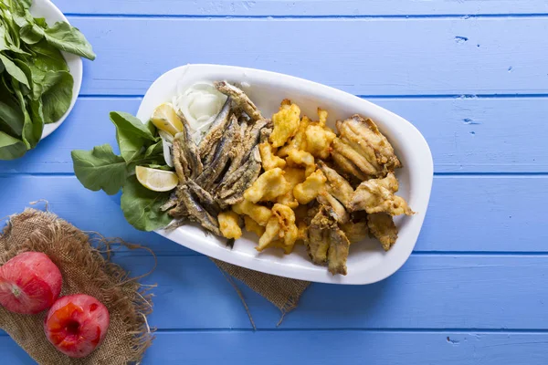 Lezzetli derin kızarmış Mix deniz ürünleri balık plaka mezgit, hamsi, sardalya, mavi rustik ahşap arka plan üzerinde beyaz soğan, limon, domates, roka salatası dilimlenmiş.