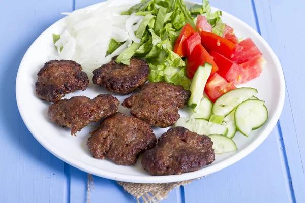 Izgara sığır eti köfte porsiyon dilimlenmiş salatalık, beyaz soğan, küp daire plakalı domates ve marul salatası mavi rustik ahşap masanın üstüne kesmek. Ekmek kırıntıları, tereyağı ve baharat gibi maddeler.