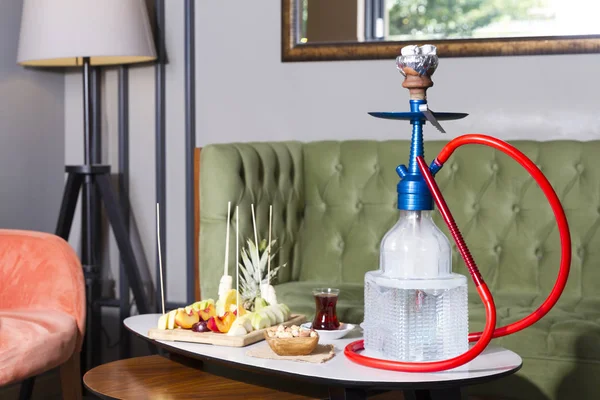Traditional Arabic and Turkish shisha, hookah or Water pipe serving with fresh fruits, hot black tea and nuts on table, modern cafe lounge design. Led light in water.