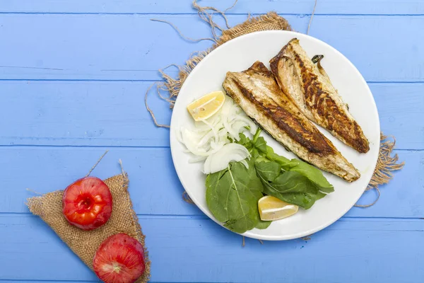 Lezzetli ızgara fileto levrek porsiyon dilimlenmiş soğan, domates, yeşil roka salatası ve sulu limon rustik mavi ahşap masa arka plan üzerinde oval beyaz plakalı füme. Alan, kopyalama kimse.