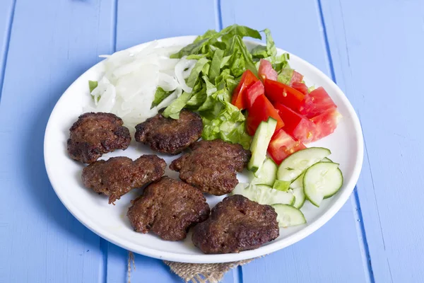 Izgara sığır eti köfte porsiyon dilimlenmiş salatalık, beyaz soğan, küp daire plakalı domates ve marul salatası mavi rustik ahşap masanın üstüne kesmek. Ekmek kırıntıları, tereyağı ve baharat gibi maddeler.