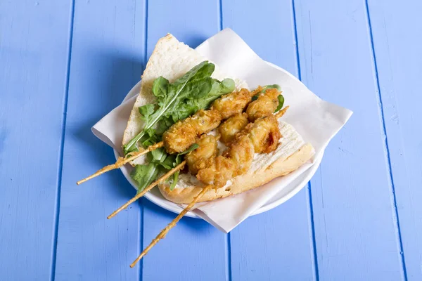 Lezzetli altın derin kızarmış pirzola midye şişleri yoğurt sos tartar ile hizmet veren sandviç, domates, dilimlenmiş beyaz soğan, maydanoz ve yeşil roka salatası mavi rustik masa arka plan üzerinde. Midye tava ekmek.