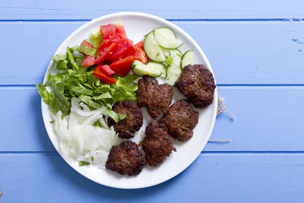 Izgara sığır eti köfte porsiyon dilimlenmiş salatalık, beyaz soğan, küp daire plakalı domates ve marul salatası mavi rustik ahşap masanın üstüne kesmek. Ekmek kırıntıları, tereyağı ve baharat gibi maddeler.
