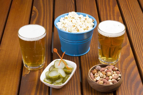 Two Glass of Draught Beer serving with snack nuts, popcorn and sliced pickles on wood table background.