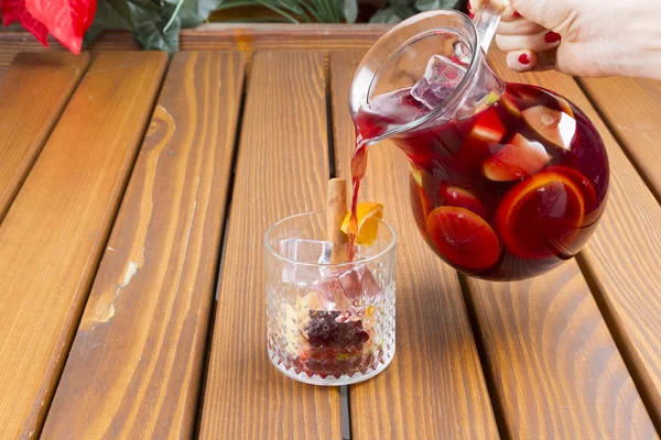 Hand pouring red wine sangria or punch with fruits and ice in glasses and pincher. Homemade refreshing fruit sangria over rustic wooden table, copy space.