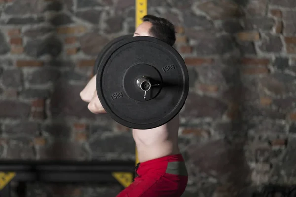 Topless male athlete practicing olympic lifts at gym with brick wall background.