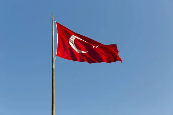 Turkish flag on long metal iron pole waving in blue sky. Red flag featuring a white star and crescent. Flag is often called al bayrak and is referred to as al sancak in the Turkish national anthem.