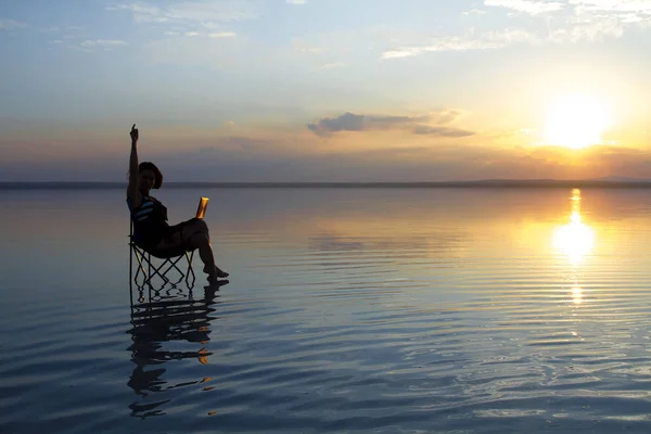 woman with laptop resting in chair at seashore during sunset