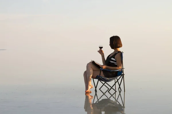 woman with glass of wine resting in chair at seashore during sunset