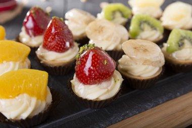 Tahta masadaki meyveli tatlıların yakın plan görüntüsü. Taze çilekli tartlar, ananas, mango, kivi, portakal ve muz ahşap masa arkasında..