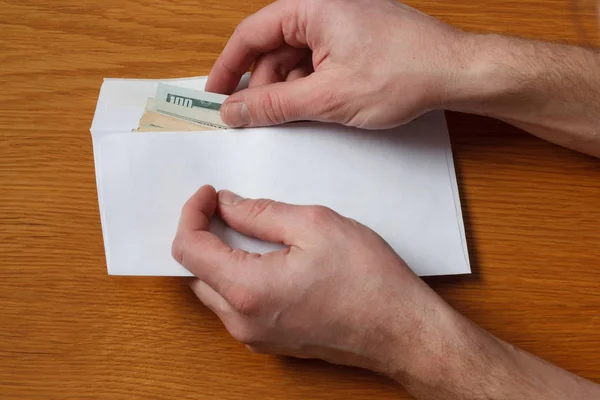 Greedy hairy man\'s hands takes a bribe of some US dollars banknotes in the blank white envelope over a wooden oak table in the office