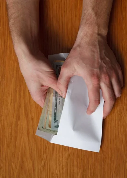 Greedy hairy man\'s hands takes a bribe of some US dollars banknotes in the blank white envelope over a wooden oak table in the office