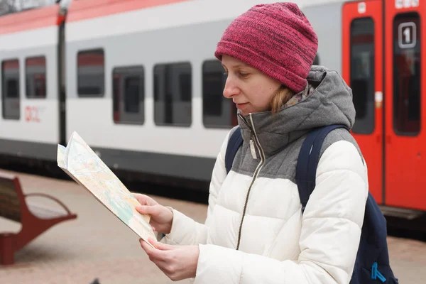 Adult young woman in warm sacual clothes travelling and looking for a route on the map how to get somewhere at railway station on the background of the train