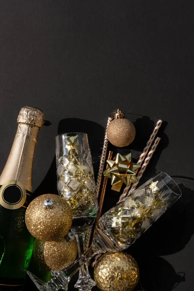 Flat lay of a bottle of champagne with crystal glasses confetti, baubles and straws on a black background — Stock Photo, Image