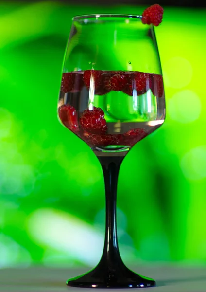 bright red raspberries in a crystal glass on a black leg inside with a sparkling drink on a bright green background of plants