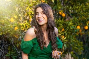 Kameraya bakarak ve gülümseyerek, bir turuncu meyve tutan güzel genç kadın, açık havada bir turuncu meyve bahçesi, günbatımında. Sağlıklı yaşam konsepti, cilt ve saç bakım kavramı.