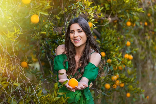 Kameraya bakarak ve gülümseyerek, bir turuncu meyve tutan güzel genç kadın, açık havada bir turuncu meyve bahçesi, günbatımında. Sağlıklı yaşam konsepti, cilt ve saç bakım kavramı.
