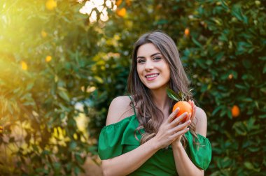 Kameraya bakarak ve gülümseyerek, bir turuncu meyve tutan güzel genç kadın, açık havada bir turuncu meyve bahçesi, günbatımında. Sağlıklı yaşam konsepti, cilt ve saç bakım kavramı.